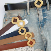 کمربند زنانه با سگک طلایی لوزیشکل در رنگهای متنوع شامل مشکی، قهوهای، سفید و سرمهای، همراه با شلوار جین در پسزمینه