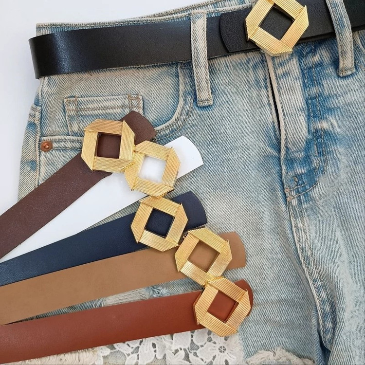 کمربند زنانه با سگک طلایی لوزیشکل در رنگهای متنوع شامل مشکی، قهوهای، سفید و سرمهای، همراه با شلوار جین در پسزمینه